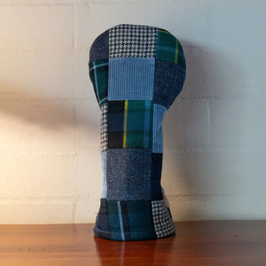 A golf club headcover made of a patchwork fabric in shades of blue, green, and black plaid and houndstooth, displayed on a wooden surface against a white brick wall.