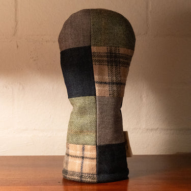 A golf club headcover made of patchwork tweed fabric in earthy green, beige plaid, and dark navy tones, resting on a wooden surface against a textured beige wall.