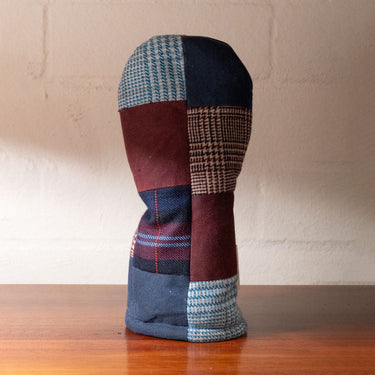 A handmade patchwork golf club headcover made from various fabric patterns including houndstooth, plaid, and solid maroon, standing on a wooden table against a brick wall.