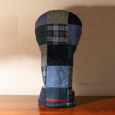 A patchwork golf club headcover made from various blue, green, gray, and beige fabrics, standing upright on a wooden surface against a beige wall.