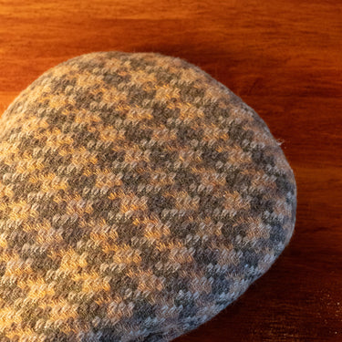 A close-up of a textured tweed flat cap in a gray and tan houndstooth pattern, resting on a warm brown wooden surface.