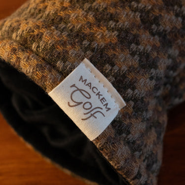 Close-up of a textured tweed knit hat in brown, beige, and black tones, with a white fabric tag reading 'MACKEM GOLF' sewn onto the side.